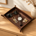 Brown leather valet tray with snap corners holding keys, a watch, sunglasses and coins on an entryway console