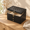 Black quilted faux leather jewelry box with two drawers on an oak dresser, lid open showing velvet lining and gold rings
